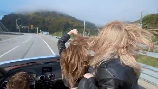 Dwa piękne młode dziewczyny jeździć w czerwony kabriolet wśród gór. Drogi na autostradzie. Ubrani w czarne skórzane kurtki. Włosów łopocze na wietrze. Ciesz się podróż i wolność. — Wideo stockowe