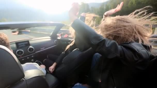 Dos hermosas jóvenes viajan en un cabriolet rojo entre las montañas. Camino en la carretera. Vestido con chaquetas de cuero negro. Cabello revoloteando en el viento. Disfruta de viajes y libertad . — Vídeo de stock