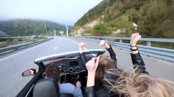 Dos hermosas jóvenes viajan en un cabriolet rojo entre las montañas. Camino en la carretera. Vestido con chaquetas de cuero negro. Cabello revoloteando en el viento. Disfruta de viajes y libertad . — Vídeo de stock