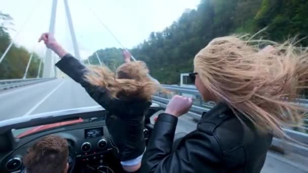 Deux belles jeunes filles chevauchent dans un cabriolet rouge au milieu des montagnes. Route sur l'autoroute. Habillé de vestes en cuir noir. Cheveux flottants dans le vent. Profitez du voyage et de la liberté . — Video