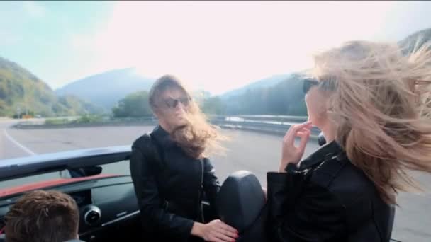 Zwei schöne junge Mädchen fahren in einem roten Cabrio durch die Berge. Straße auf der Autobahn. bekleidet mit schwarzen Lederjacken. Haare flattern im Wind. Reisen und Freiheit genießen. — Stockvideo