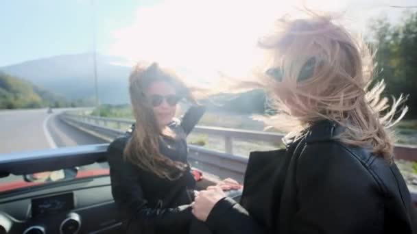 Duas lindas garotas montam em um cabriolet vermelho entre as montanhas. Estrada na auto-estrada. Vestido com jaquetas de couro preto. Cabelo a tremer ao vento. Desfrute de viagens e liberdade . — Vídeo de Stock