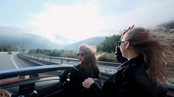 Two beautiful young girls ride in a red cabriolet among the mountains. Road on the highway. Dressed in black leather jackets. Hair fluttering in the wind. Enjoy travel and freedom. — Stock Video
