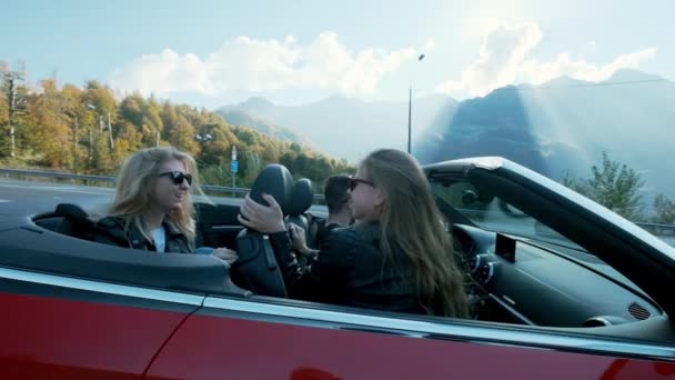 Привлекательные европейские подруги поют, танцуют и хлопают в ладоши за рулем роскошного открытого автомобиля вдоль высоких гор — стоковое видео
