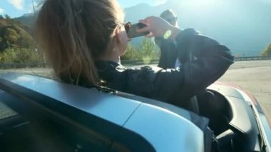 İki genç güzel kız siyah gözlüklü ve deri ceketler kırmızı Cabriolet yüksek dağlara yakın. Onlar bir fotoğraf oturumu yaptığınız, kendilerini bir smartphone cep telefonu ile fotoğraf çekmek, iyi eğlenceler.