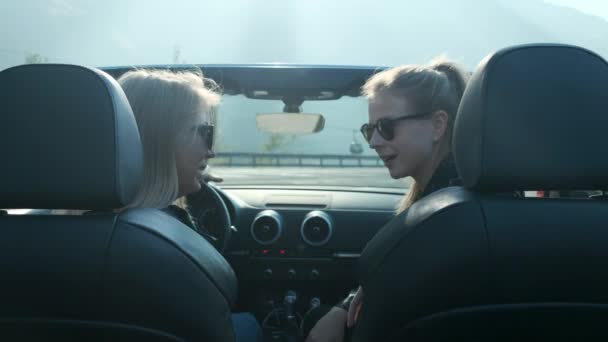 Två unga flickor i en cabriolet nära de höga bergen. Prata bakom ratten. flicka korrigerar bil backspegeln — Stockvideo