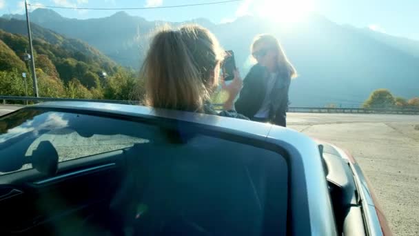 Dos chicas hermosas jóvenes en gafas negras y chaquetas de cuero en un cabriolet rojo cerca de las altas montañas. Hacen una sesión de fotos, se toman fotos en un teléfono inteligente, se divierten . — Vídeo de stock