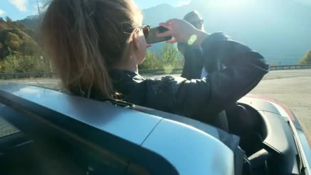 Dos chicas hermosas jóvenes en gafas negras y chaquetas de cuero en un cabriolet rojo cerca de las altas montañas. Hacen una sesión de fotos, se toman fotos en un teléfono inteligente, se divierten . — Vídeo de stock