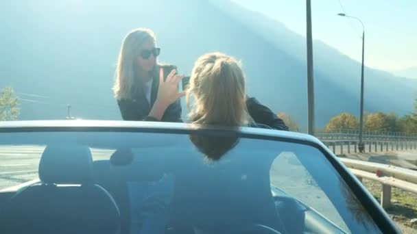 Dos chicas hermosas jóvenes en gafas negras y chaquetas de cuero en un cabriolet rojo cerca de las altas montañas. Hacen una sesión de fotos, se toman fotos en un teléfono inteligente, se divierten . — Vídeos de Stock