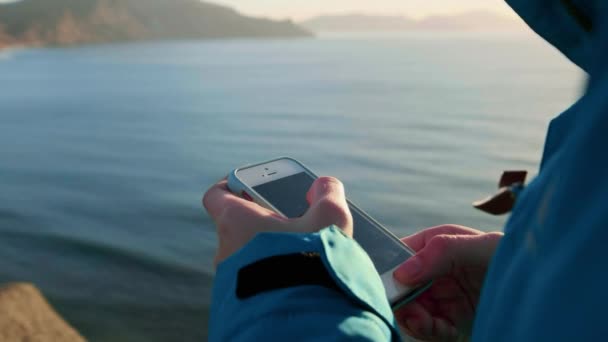Ein junges attraktives Mädchen benutzt ein Smartphone. schreibt Nachrichten oder schickt ein Foto in soziale Netzwerke. genießt das Meer im Urlaub. Es steht auf einer Klippe bei Sonnenuntergang oder Sonnenaufgang. mit blauer Jacke und rotem Hut. — Stockvideo