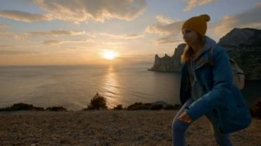 Ağır çekim, Close Up: akşam güneşi güneşlenme uçurumun kenarında duran ve yüksek rocky Dağları karşı ellerini yükselterek genç erkek. Mutlu kız zevk başarı ve çarpıcı görünümü