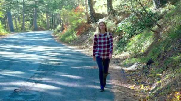 Portrait: Traveler woman hitchhiking on a forest road and walking. Young happy backpacker woman looking for a ride to start a journey on a sunlit country road — Stock Video