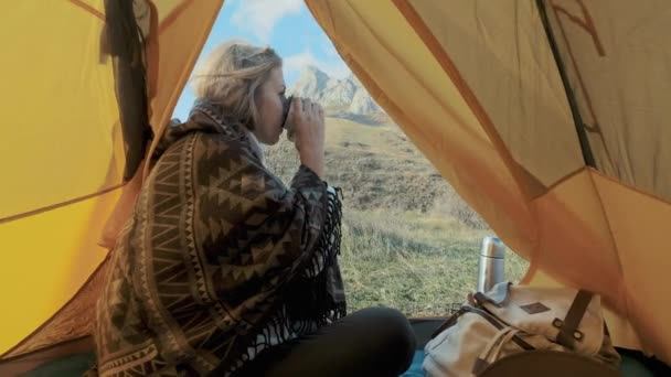 Ελκυστική κοπέλα πεζοπορία κάθεται κοντά στην είσοδο στη σκηνή στο φόντο των βουνών. Πίνοντας καφέ ή τσάι από μια κούπα. Απολαμβάνει τις διακοπές. Η έννοια της ελευθερίας στα βουνά. Κριμαία — Αρχείο Βίντεο