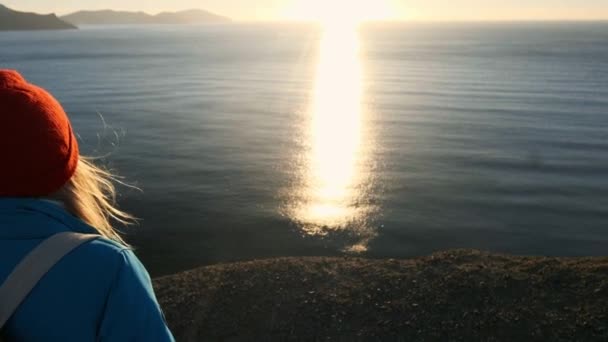 Jovem atraente menina caminhante vai ao longo das bordas arenosas ou penhasco à beira-mar. Levanta as mãos, olha para o nascer do sol dourado ou para o pôr do sol. Goza de uma sensação de sucesso. Sobe em uma montanha alta e bonita . — Vídeo de Stock