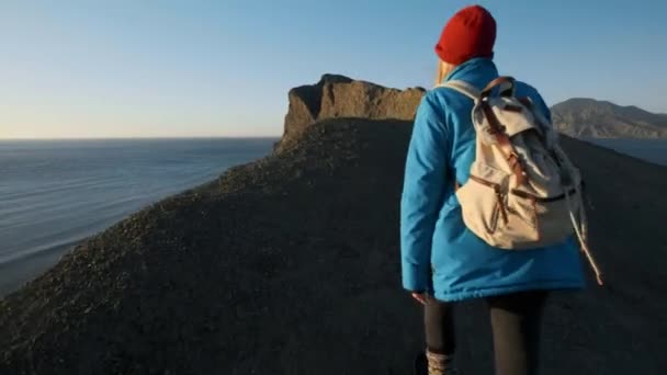 Young attractive girl hiker goes along the sandy ledges or seaside cliff. Raises his hands up, looks at the golden sunrise or sunset. Enjoys a sense of success. Climbs on a high beautiful mountain. — Stock Video