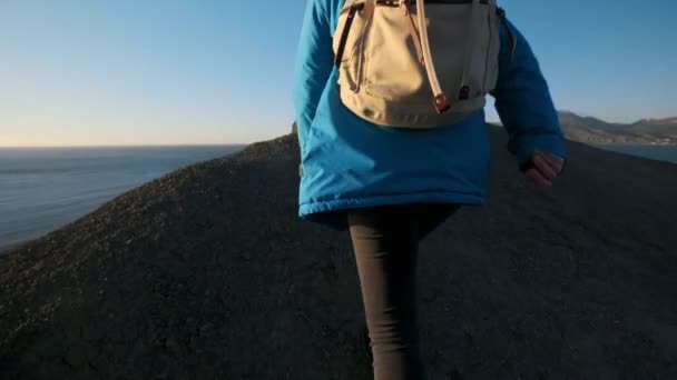 Junge attraktive Wanderin geht entlang der sandigen Felsvorsprünge oder Klippen am Meer. hebt die Hände, blickt auf den goldenen Sonnenaufgang oder Sonnenuntergang. Ein Erfolgserlebnis. klettert auf einen hohen, schönen Berg. — Stockvideo