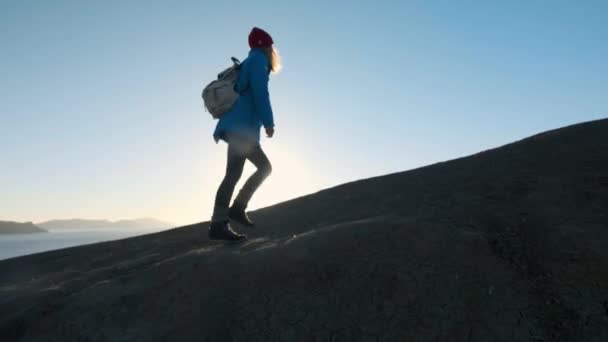 年轻迷人的女孩徒步者沿着沙质的边缘或海边的悬崖。举起双手, 看着金色的日出或日落。享受成功的感觉。爬上美丽的高山. — 图库视频影像