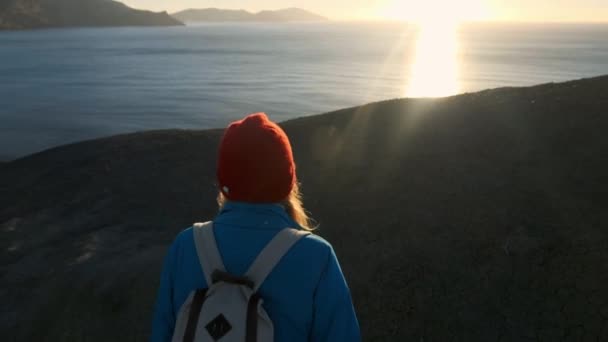 Young attractive girl hiker goes along the sandy ledges or seaside cliff. Raises his hands up, looks at the golden sunrise or sunset. Enjoys a sense of success. Climbs on a high beautiful mountain. — Stock Video
