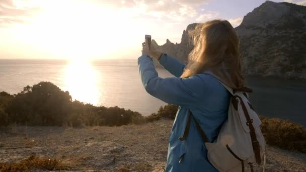 Молодая привлекательная женщина наблюдая закат солнца или восход солнца над заливом с вершины горы, девушка делает панорамные фотографии на телефоне. Портативный атмосферный выстрел. Крым, Украина — стоковое видео