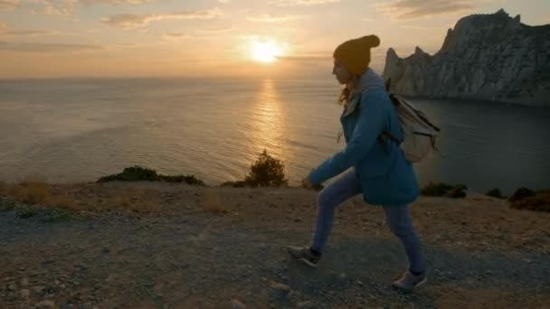 Slowmotion, Close Up: Jonge vrouw staande op de rand van de klif en verhogen haar handen tegen hoge rocky bergen zonnebaden in de avond zon. Gelukkig meisje genieten van succes en prachtig uitzicht — Stockvideo