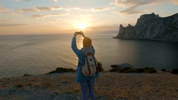 Giovane donna attraente che guarda il tramonto o l'alba sulla baia dalla cima della montagna, ragazza prende foto panoramiche sul telefono.Capelli nel vento. Colpo atmosferico palmare. Crimea, Ucraina — Video Stock