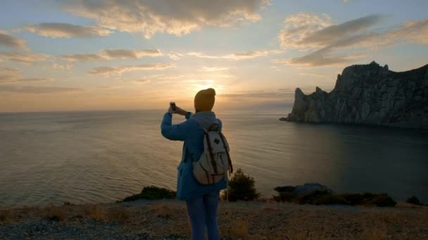 Ung vacker flicka vandring gör foto på smartphone. Skjuter en vacker solnedgång på en udde vid havet. Nått målet. Krim, Ukraina. Begreppet frihet och enhet med naturen. — Stockvideo