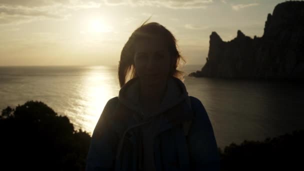 Close up retrato da jovem mulher sorrindo com o cabelo vermelho soprando no vento olhando para o pôr do sol sobre o oceano Menina vestindo capa de chuva amarela trekking na Escócia Slow Motion — Vídeo de Stock