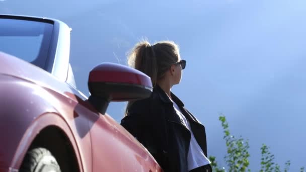 Una joven morena con estilo en una chaqueta de cuero y gafas de sol está de pie junto a un convertible rojo — Vídeos de Stock