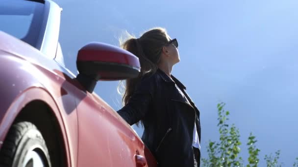Een brunette meisje in stijlvol in een leren jas en zonnebril staat naast een rode Cabrio — Stockvideo