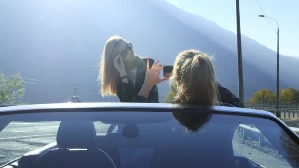 Two beautiful young stylish girls take photos on a smartphone inside a red convertible. Funny photo shoot on vacation. Posing and enjoying the rest among the mountains. — Stock Video