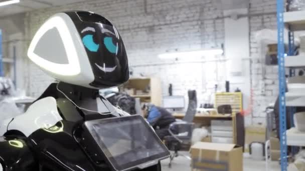 Ένα νέο ρομπότ στέκεται σε ένα δωμάτιο στο υπερώο. Εργοστάσιο για την παραγωγή των ρομπότ. Το ρομπότ αναβοσβήνει και χαμόγελα. — Αρχείο Βίντεο