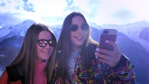 Dos chicas jóvenes suben a las montañas, hacen selfies en el fondo, sonriendo. Disfruta de las vacaciones de montaña de invierno. Diviértete en la estación de esquí de invierno. Día soleado, detrás de la montaña — Vídeos de Stock