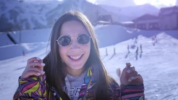 Encantadora jovencita sonriendo en gafas de sol en invierno. Disfruta del resto de la estación de esquí de invierno en las montañas. Mirando juguetonamente la cámara. Clima soleado, la chica está vestida con ropa de esquí . — Vídeo de stock
