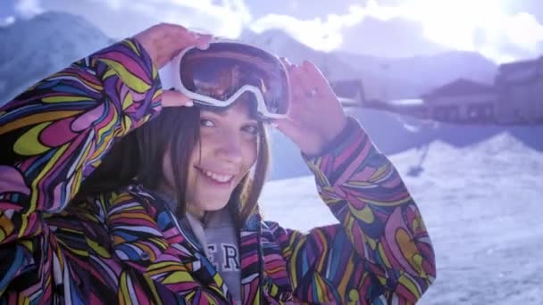 Hermosa joven con gafas de esquí. Sonríe y obtiene placer de las vacaciones de invierno. Listo para esquiar o snowboard. Vacaciones de invierno en un resort de snowboard. Situado en la cima de la montaña . — Vídeos de Stock