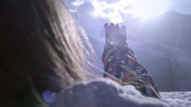 Ένα νεαρό κορίτσι κοιτάζει τον ήλιο μέσα από το χέρι και τα δάχτυλά της. Παίζοντας με τις ακτίνες του ήλιου. Απολαμβάνει τις χειμερινές διακοπές του βουνού. Ηλιόλουστη μέρα, με φόντο τα βουνά. — Αρχείο Βίντεο