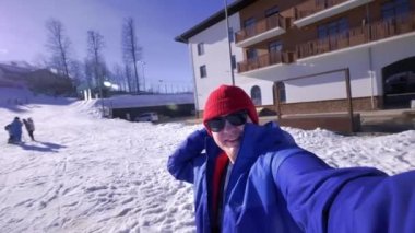 Genç bir adam kamera bakıyor, kafasına bir mızrak koyar. Kış karla kaplı dağlara sahiptir. Mavi bir kayak ceketi, kırmızı şapka ve güneş gözlüğü giyiyor. Dağlarda kış tatil