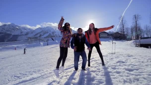 Gezelschap van vrienden die plezier hebben op vakantie in de winter bergen. Een man en twee meisjes genieten van de winter in het skigebied. Ze lopen in de sneeuw, springen. Sneeuw vliegt in de camera. Achter de berg — Stockvideo