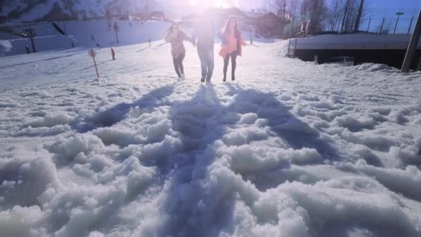 Compañía de amigos divirtiéndose de vacaciones en las montañas de invierno. Un chico y dos chicas disfrutan del invierno en la estación de esquí. Corren en la nieve, saltan. La nieve vuela hacia la cámara. Detrás del monte — Vídeos de Stock