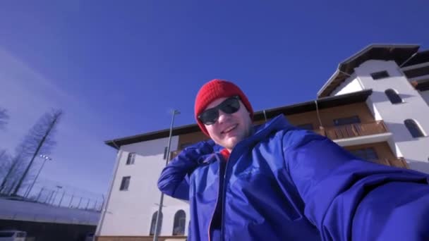 Egy fiatal srác nézi a kamerát, teszi a lándzsát a fején. Élvezi téli hófödte hegyek. Visel kék sí kabát és egy piros kalap és napszemüveg. Téli üdülőhely a hegyekben — Stock videók