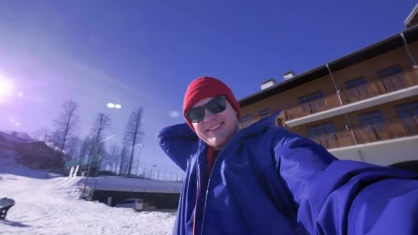 Un joven mira a la cámara, se pone una lanza en la cabeza. Disfruta de las montañas nevadas de invierno. Llevar una chaqueta de esquí azul y un sombrero rojo y gafas de sol. Vacaciones de invierno en las montañas — Vídeo de stock