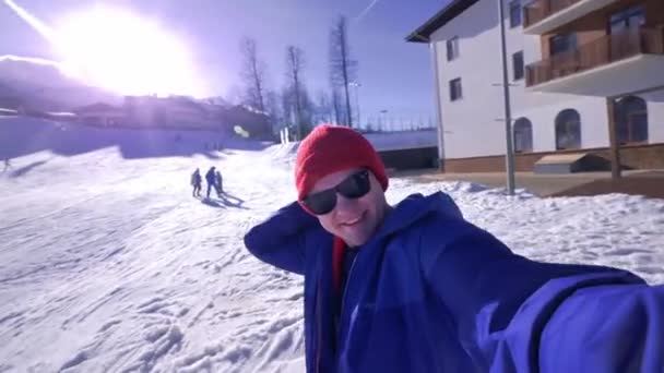 Ένας νεαρός κοιτάζει την κάμερα, βάζει ένα δόρυ στο κεφάλι του. Απολαμβάνει το χειμερινό χιονισμένο βουνό. Φορώντας ένα μπλε μπουφάν του σκι και ένα κόκκινο καπέλο και γυαλιά ηλίου. Χειμερινό θέρετρο στα βουνά — Αρχείο Βίντεο