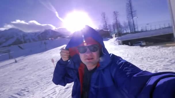 Um jovem olha para a câmara, coloca uma lança na cabeça. Gosta de montanhas cobertas de neve de inverno. Vestindo um casaco de esqui azul e um chapéu vermelho e óculos de sol. Resort de inverno nas montanhas — Vídeo de Stock