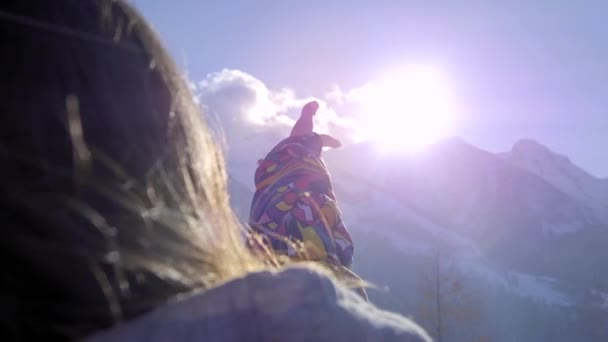 Ein liebes Mädchen blickt durch ihre Hand in die Sonne. spielt im Winter Palme in der Sonne. Im Urlaub genießt sie die schneebedeckten Berge im Winter. bekleidet mit einer farbigen Skijacke. Wintersportort in den Bergen. — Stockvideo