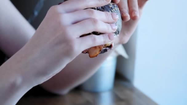 A lovely young girl has a huge burger inside a cafe. Widely opens his mouth, bites off a piece of burger. Street food, unhealthy junk food. Obesity and healthy eating problems. Slow motion, Clouse-up — Stock Video