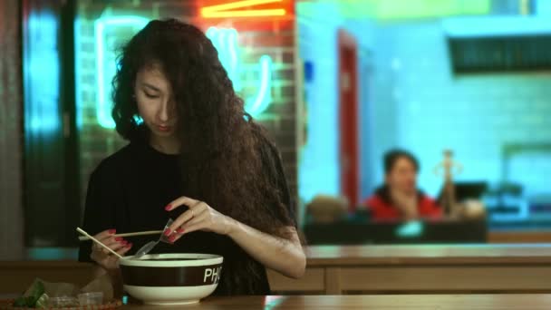 Ένα όμορφο νεαρό ασιατικό κορίτσι τρώει φο σούπα σε ένα ασιατικό καφέ. Παίρνει νουντλς με κινέζικα ξυλάκια. Κινέζικο, βιετναμέζικο ή γιαπωνέζικο καφέ ή εστιατόριο. Προσθέτει σάλτσα στη σούπα. — Αρχείο Βίντεο