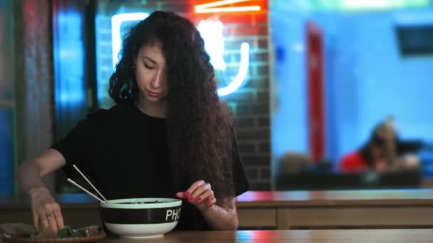 Uma linda jovem asiática está comendo sopa de pho em um café asiático. Toma macarrão com pauzinhos chineses. Café ou restaurante chinês, vietnamita ou japonês. Adiciona verduras e limão à sopa . — Vídeo de Stock