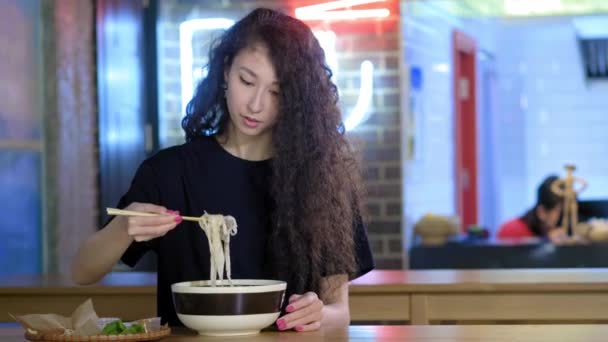 Een mooie jonge Aziatische meisje is het eten van Pho soep in een Aziatisch Cafe. Neemt noedels met Chinese eetstokjes. Chinees, Vietnamees of Japans café of restaurant. — Stockvideo