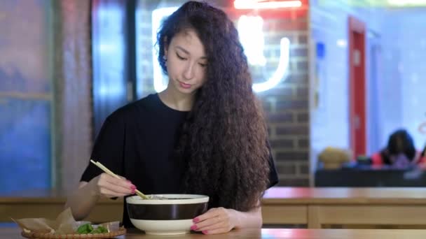 Ένα όμορφο νεαρό ασιατικό κορίτσι τρώει φο σούπα σε ένα ασιατικό καφέ. Παίρνει νουντλς με κινέζικα ξυλάκια. Κινέζικο, βιετναμέζικο ή γιαπωνέζικο καφέ ή εστιατόριο. — Αρχείο Βίντεο