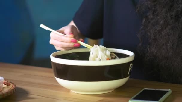 Una encantadora joven asiática está comiendo sopa pho en un café asiático. Toma fideos con palillos chinos. Café o restaurante chino, vietnamita o japonés . — Vídeo de stock