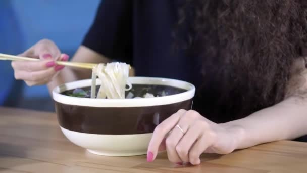 Una encantadora joven asiática está comiendo sopa pho en un café asiático. Toma fideos con palillos chinos. Café o restaurante chino, vietnamita o japonés . — Vídeo de stock